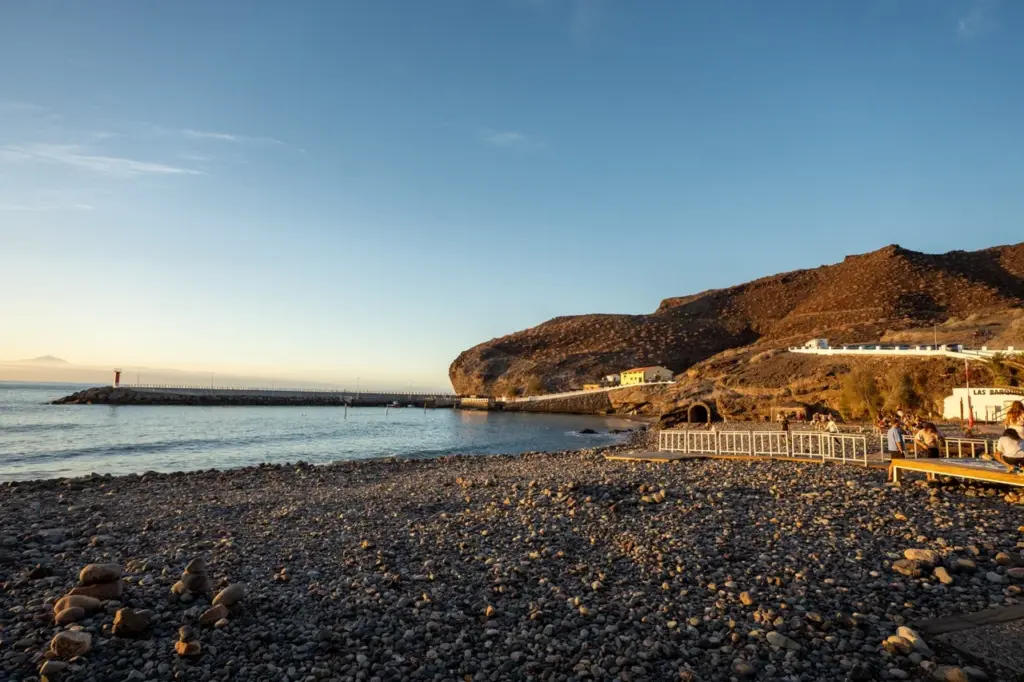 Der felsige Strand von Gran Canaria, das ruhige Meer, eine weit entfernte Mole und sonnenbeschienene Klippen unter blauem Himmel.