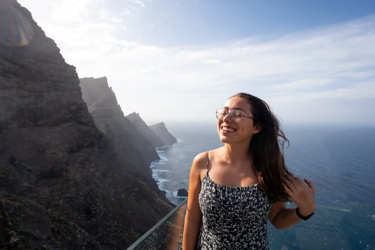 Clara in einem Kleid steht vor den Klippen Gran Canarias, mit Blick auf das Meer und die felsigen Berge im Hintergrund.