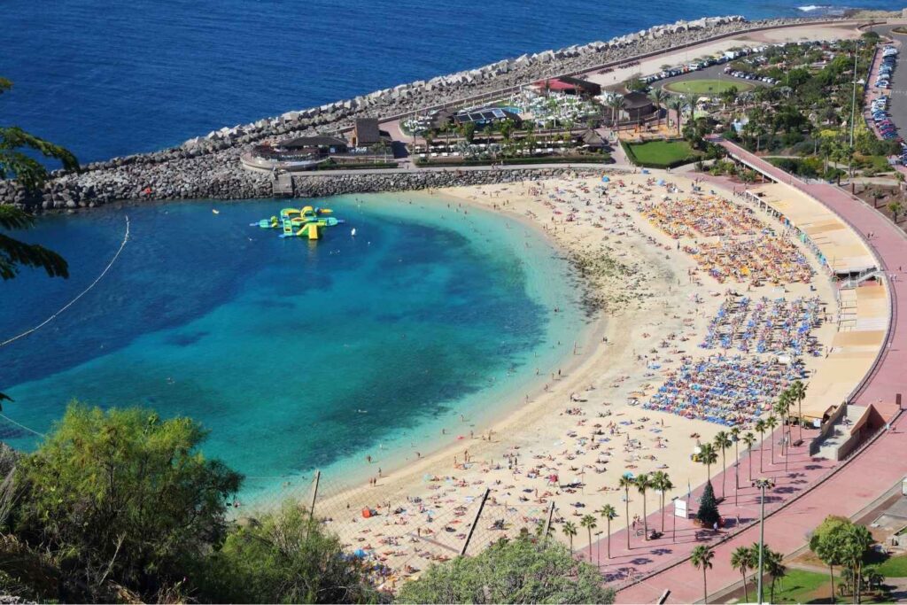 Luftaufnahme von Playa Anfi de Mar, Gran Canaria, mit goldenem Sand, blauem Meer, Sonnenanbetern und Wasserpark.