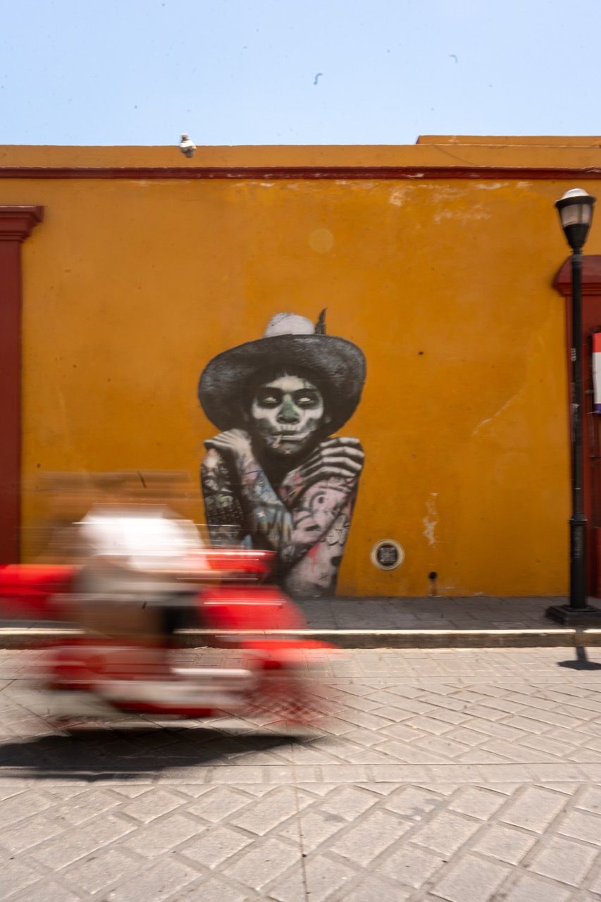 Catrina-Wandbild an oranger Hauswand in Oaxaca, davor ein fahrendes Mototaxi
