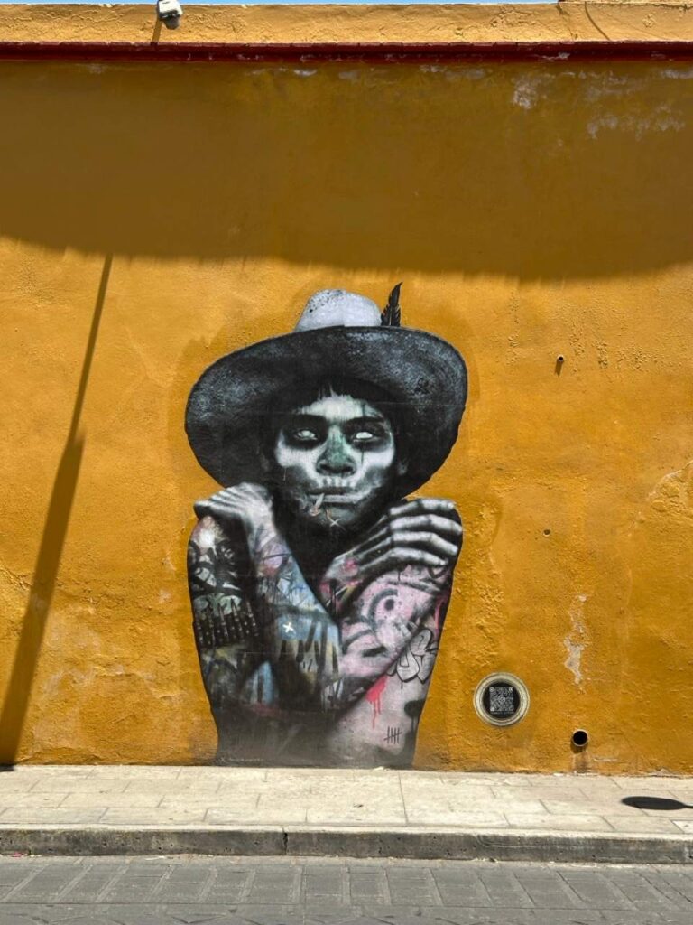 Streetart-Mural einer Skelettfigur mit Hut an ockergelber Wand in Oaxaca