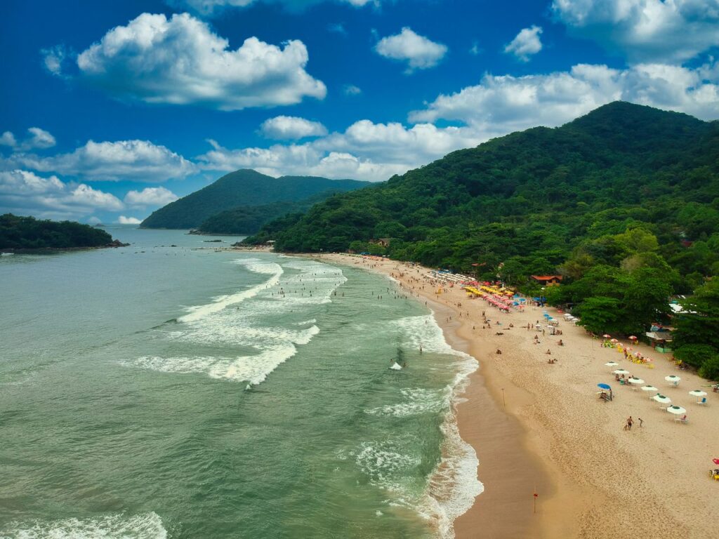 Drone view of sea with foamy waves running on sandy beach with tourists under blue sky with clouds in summer day
