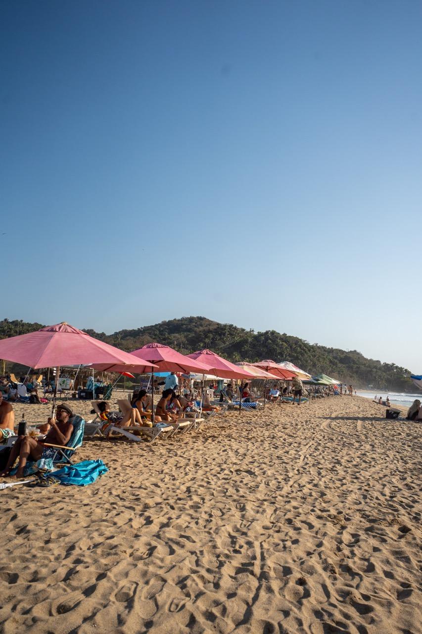 Pinke Sonnenschirme und Liegen am Strand von Sayulita mit Hügel im Hintergrund