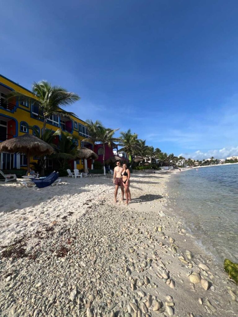 Paar am Strand vor einem bunten Strandhotel mit Palmen in Tulum