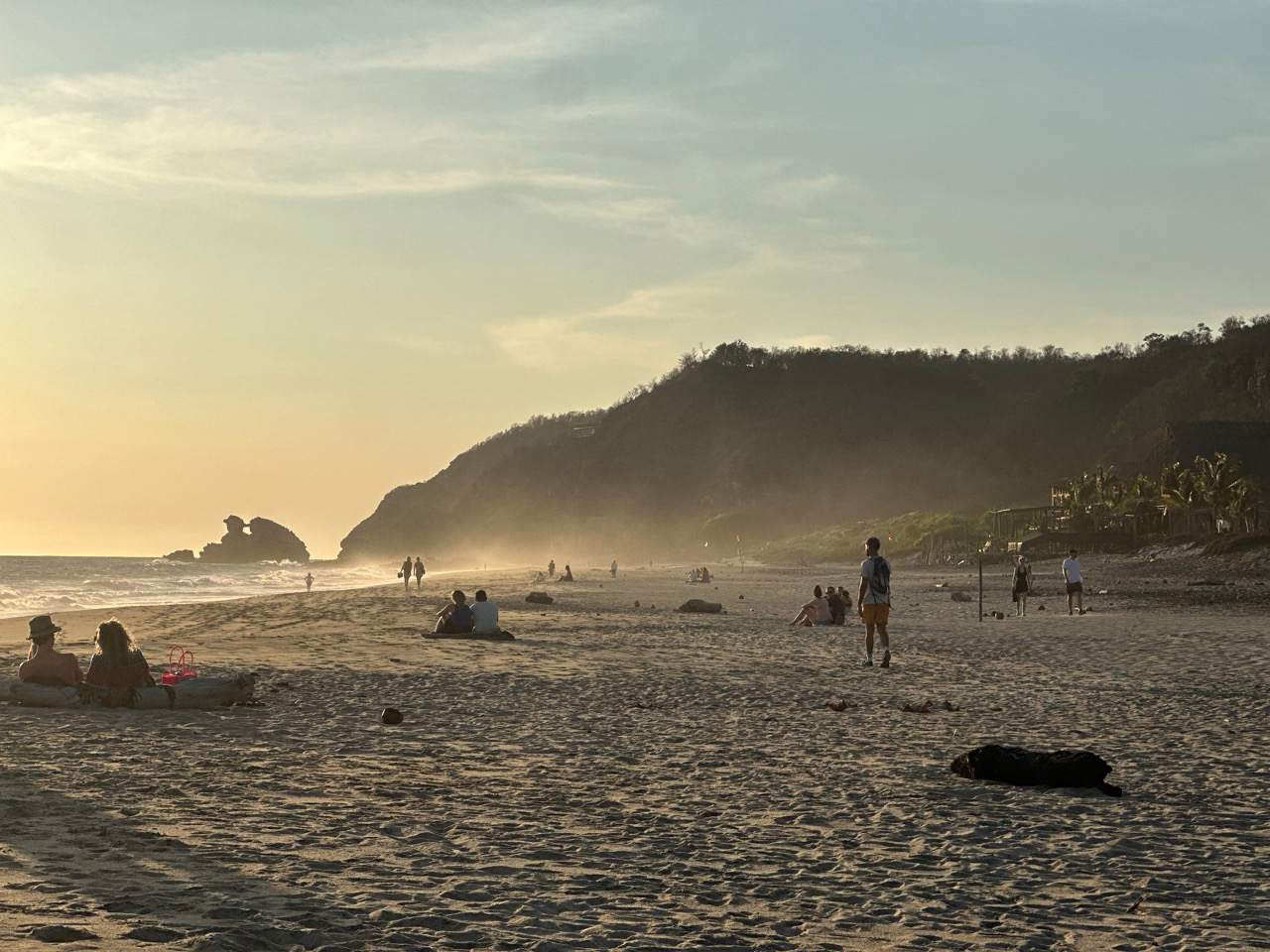 Menschen am Strand von Zipolite im goldenen Abendlicht mit Huegeln und Felsen im Dunst
