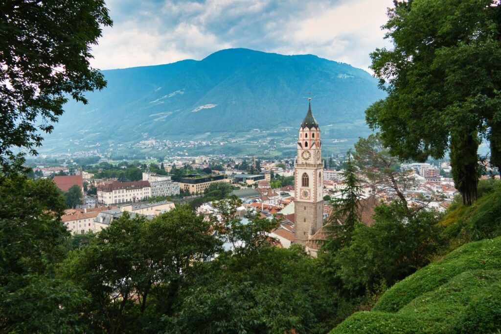 A picturesque townscape of Meran, Italy, with the iconic St. Nicholas Church tower amid lush greenery.