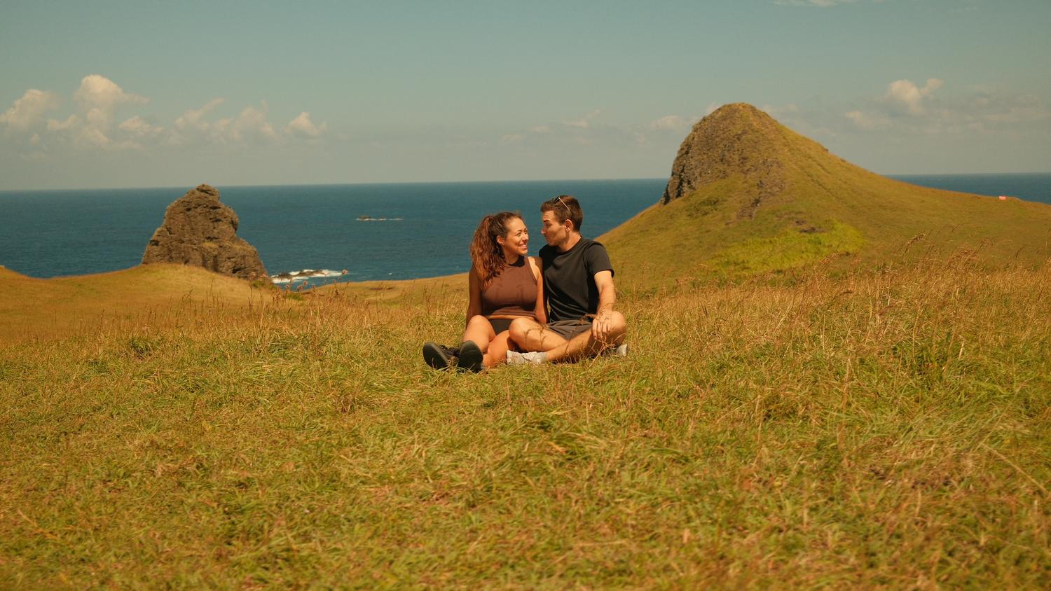 Clara und Jonathan sitzen im Gras vor Felsspitze und Meer auf Green Island